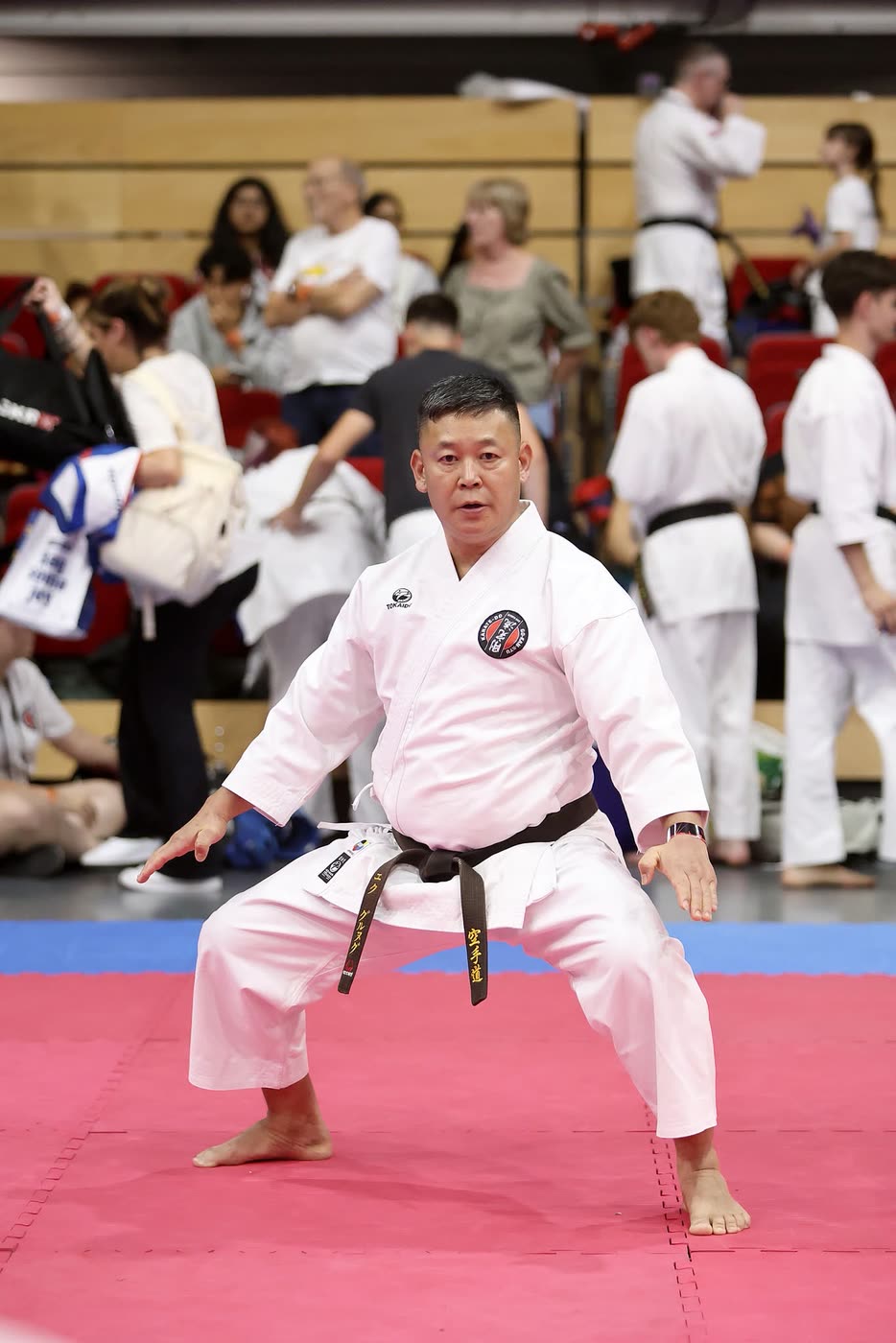 A black-belt competitor holding a low kata stance, arms extended, mid-form.